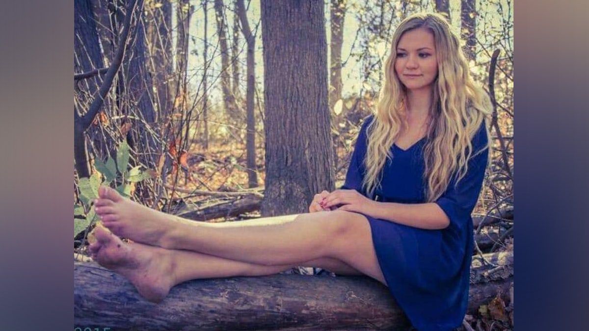 Gina draagt een blauwe jurk en zit op een boomstam in het bos.
