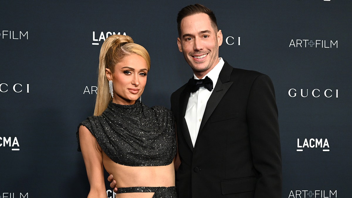 Paris Hilton in een fonkelende uitgesneden jurk met opgestoken haar poseert met echtgenoot Carter Reum in een klassieke smoking op het tapijt