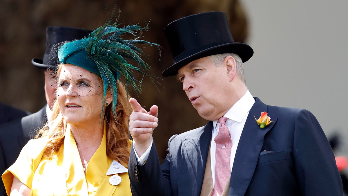 Prins Andrew in een donkerblauw pak en zwarte hoge hoed wijst naar boven terwijl Sarah Ferguson, in een gele jurk en groene fascinator, toekijkt.