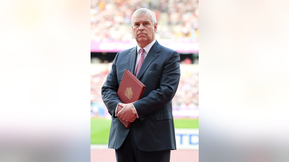 Prins Andrew draagt een donker pak en rode stropdas en houdt een bordeauxrood boek vast.