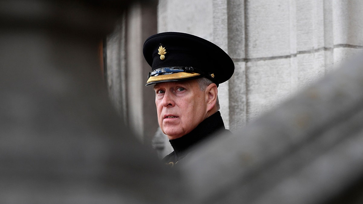 Prins Andrew draagt een militair uniform en hoed en kijkt bedroefd achter een grijs gebouw.